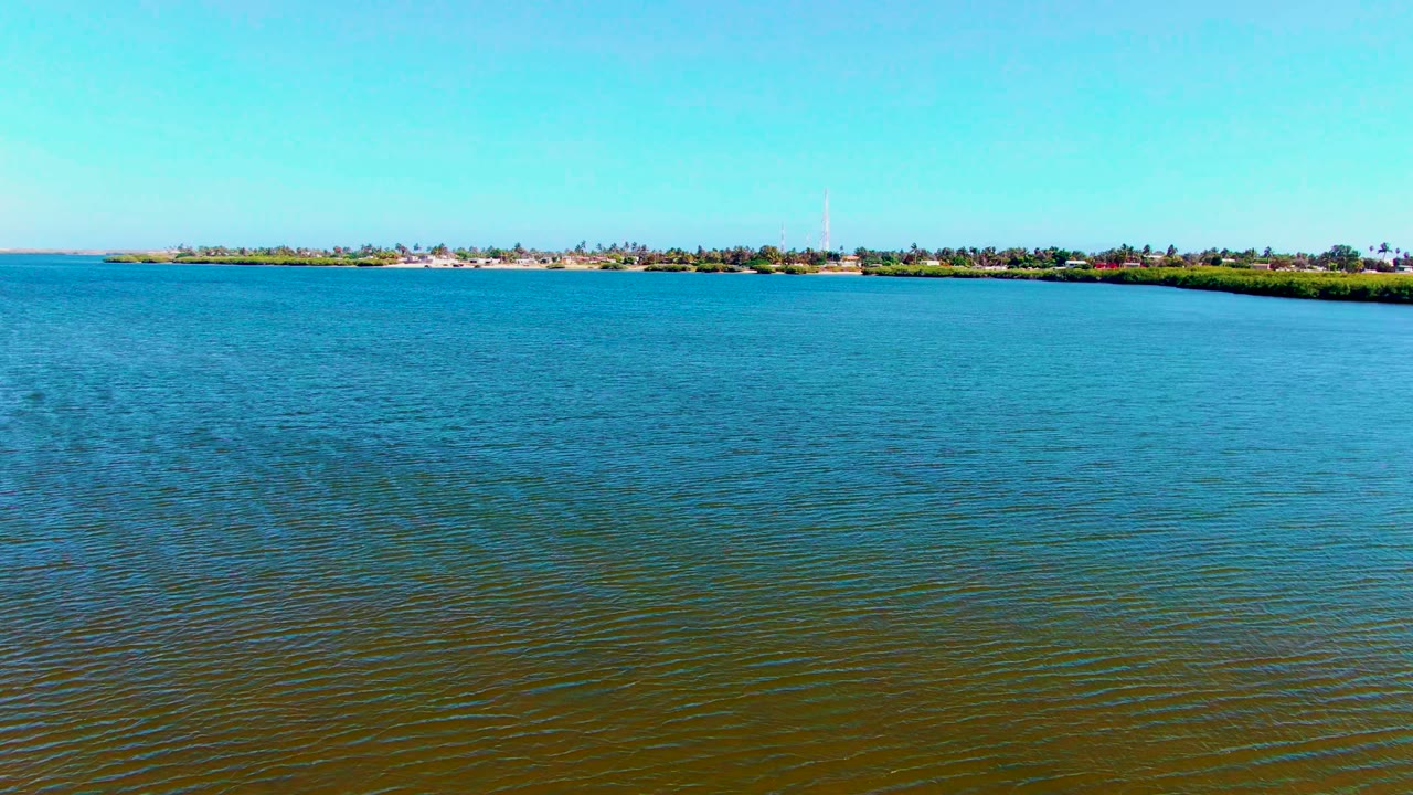 Opening frame of calm rippled water in San Carlos, Baja California Sur, with a low distant shoreline, trees, small buildings, a tall tower-like structure, and no visible people.