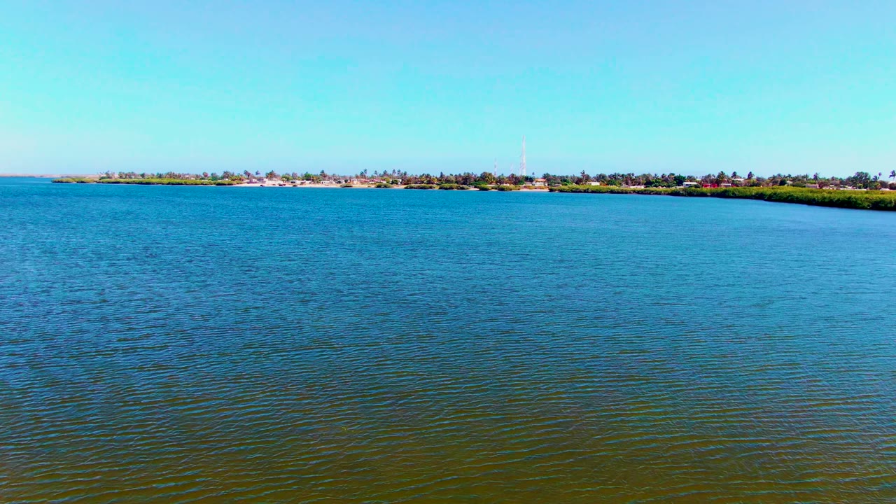 Third frame of the San Carlos shoreline across blue-green water, with small buildings and a tall tower-like structure visible along the thin background land strip, with no visible people.
