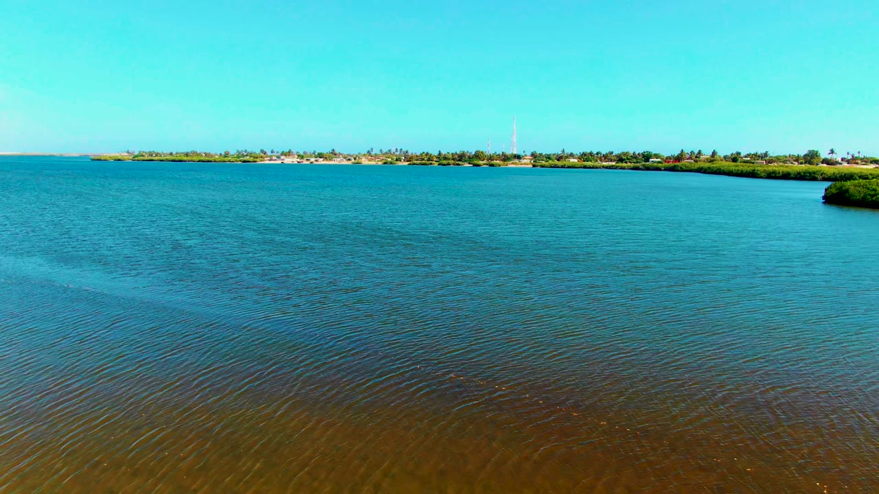 San Carlos, Baja California Sur aerial frame with open blue water dominating the view, a small mangrove edge at right, distant low shore buildings, a tower, and no visible people.