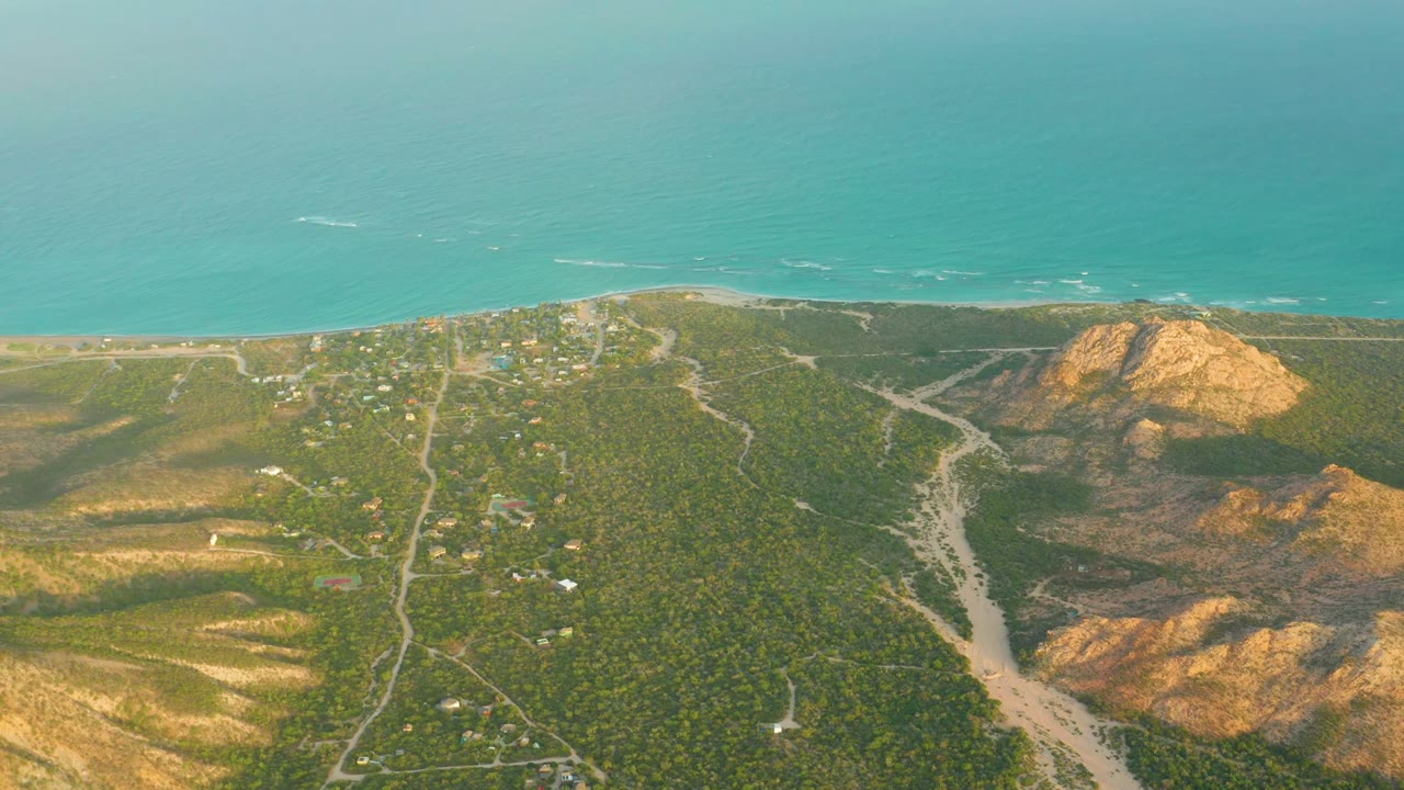 Aerial view of Cabo Pulmo BCS Mexico with turquoise ocean above a sandy shore, winding dirt roads through green vegetation, ochre hills at right, and no visible people.