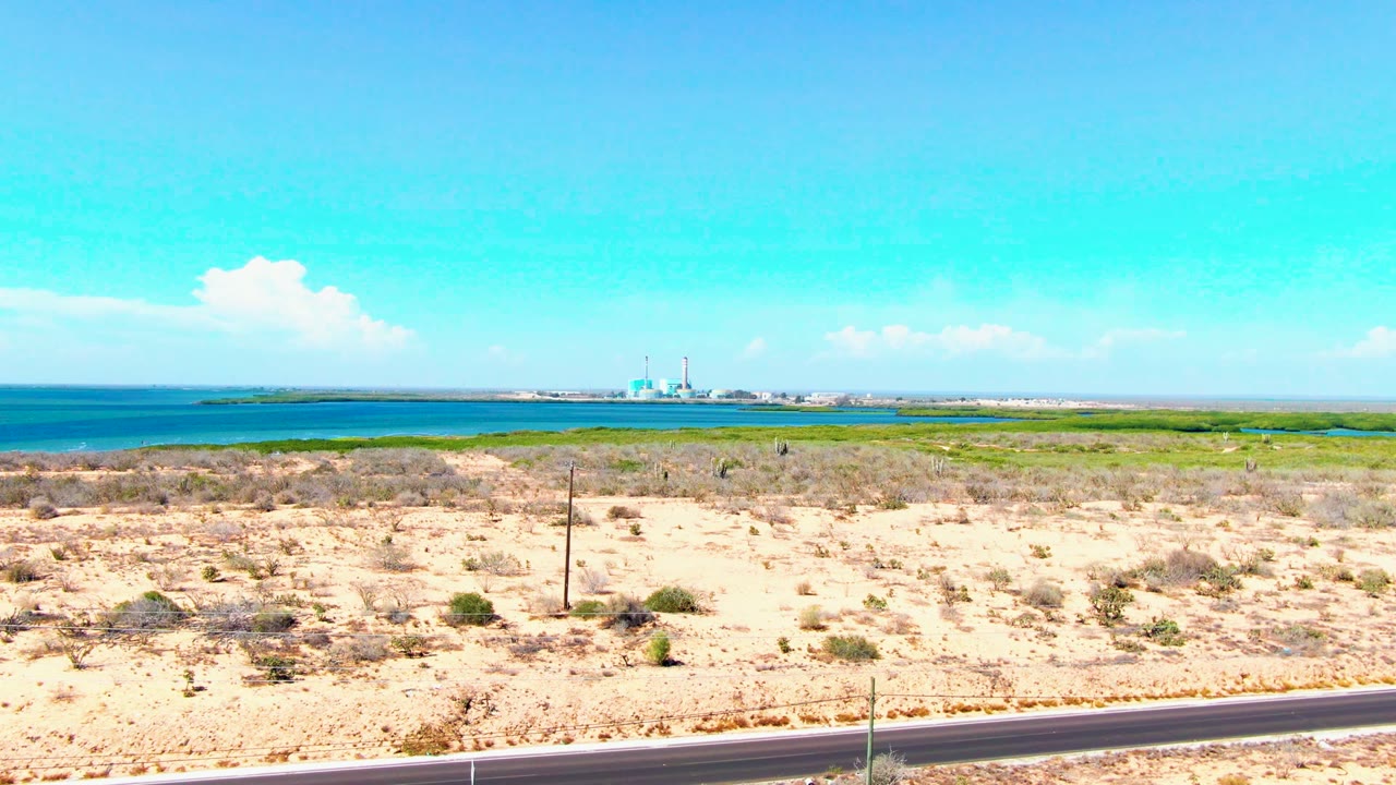 San Carlos, Baja California Sur coastal desert view with a paved road in the foreground, blue ocean beyond sandy scrub, a distant dome and minaret, and no visible people.
