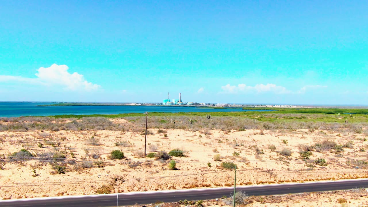 A wide San Carlos, Baja California Sur view shows road edge, sparse desert plants, blue coastal water, distant waterfront buildings with a dome and minaret, and no visible people.