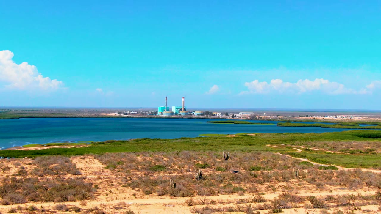 Wide elevated view in San Carlos, Baja California Sur, Mexico, showing sandy scrubland, blue water, green shoreline vegetation, distant industrial towers, and a small person far from camera.