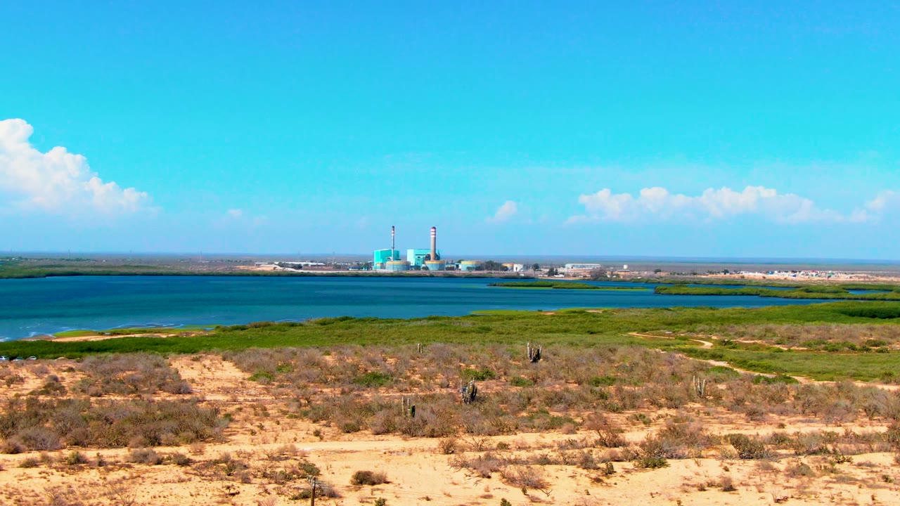A broad San Carlos, Baja California Sur view shows tan open ground, scattered brush, green vegetation along blue water, industrial stacks on the horizon, and a very small person walking away.
