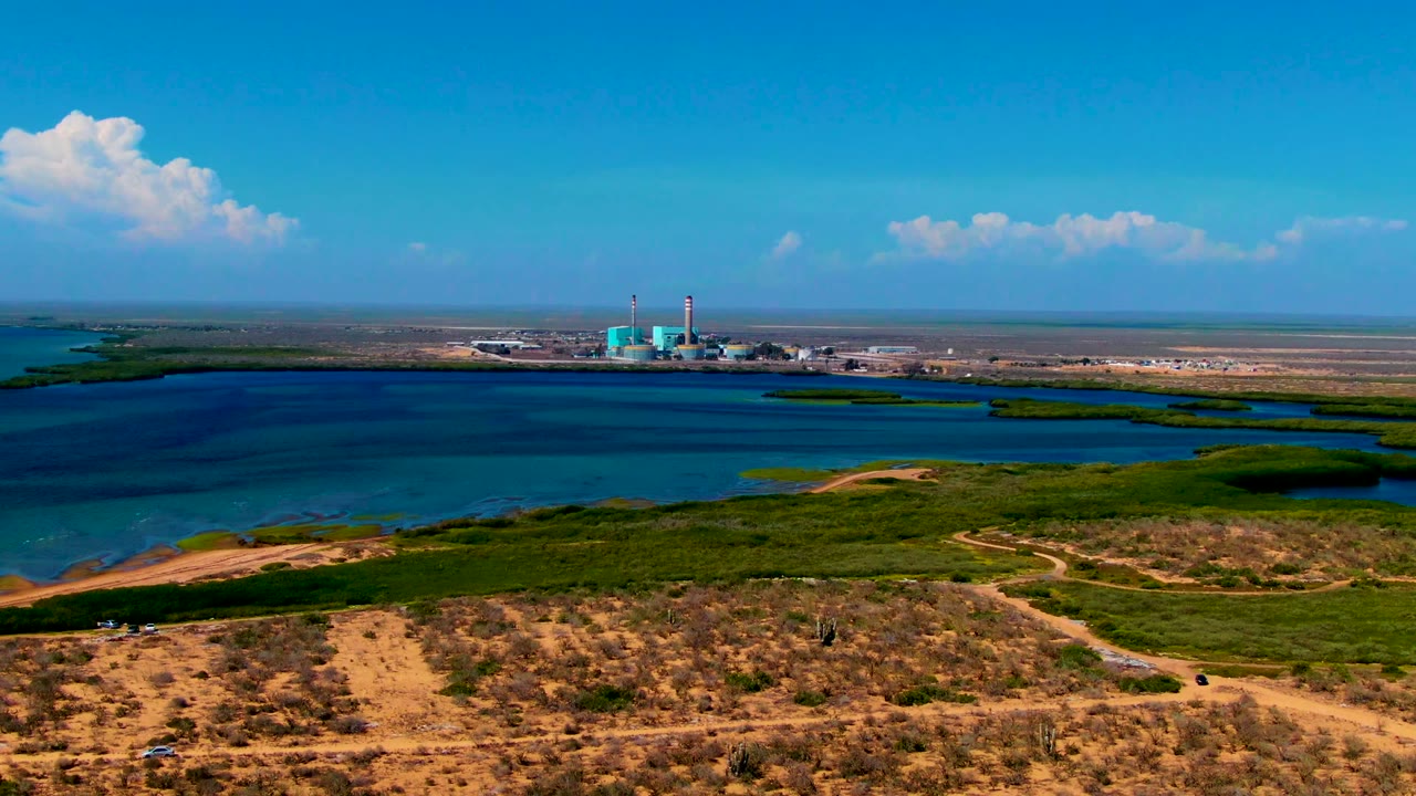 Opening aerial frame in San Carlos, Baja California Sur, showing sandy scrub dunes, a broad blue waterway, an industrial plant with two stacks on the far shore, scattered clouds, and no visible people.