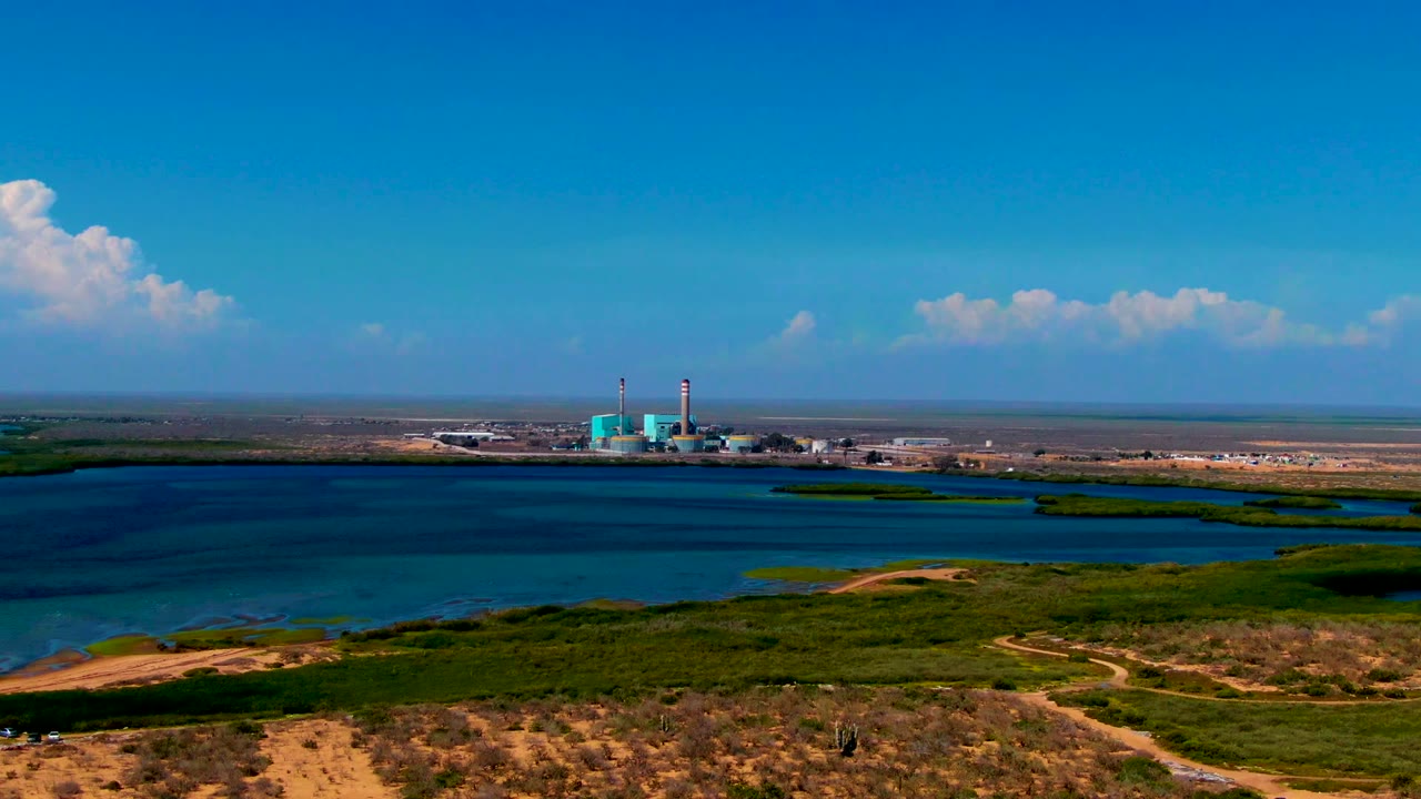 Wide San Carlos, Baja California Sur aerial frame showing the industrial complex farther to the right across the water, dry dunes and green patches below, a low horizon, and no visible people.