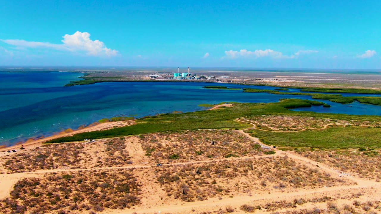 Aerial view in San Carlos, Baja California Sur showing sandy scrubland, green shoreline vegetation, blue coastal water, a distant industrial structure, bright sky, and no visible people.