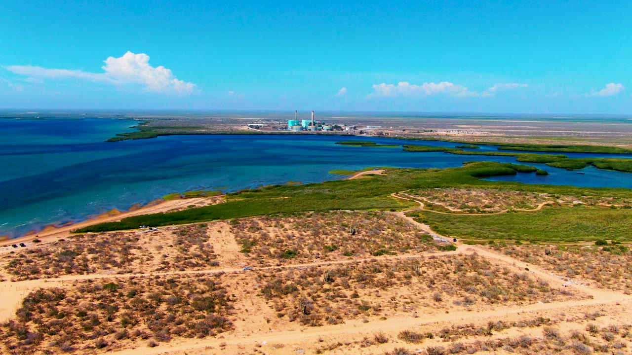 Drone-height view over sandy coastal terrain toward blue water and industrial buildings.