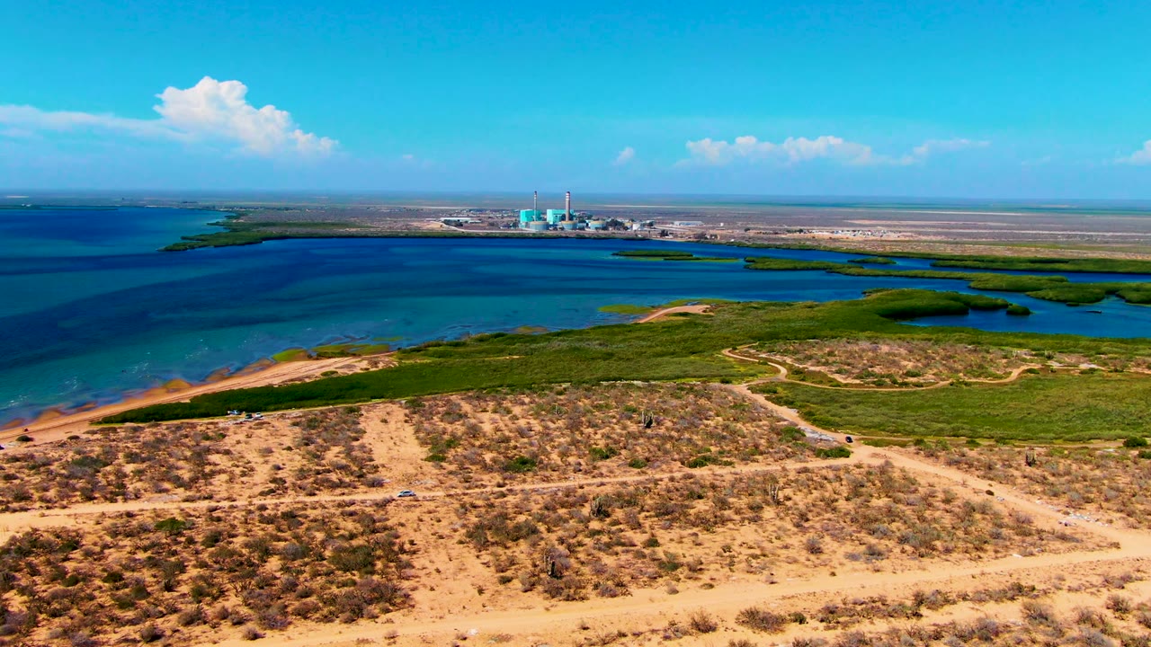 Wide aerial frame in San Carlos, Baja California Sur with dry desert foreground, green wetland bands, blue bay water, a small industrial site on the far shore, and no visible people.