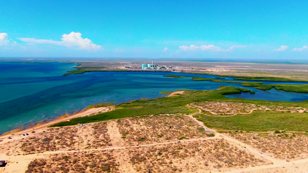 High coastal view with scrub dunes, blue-green water, wetlands, clouds, and horizon structures.