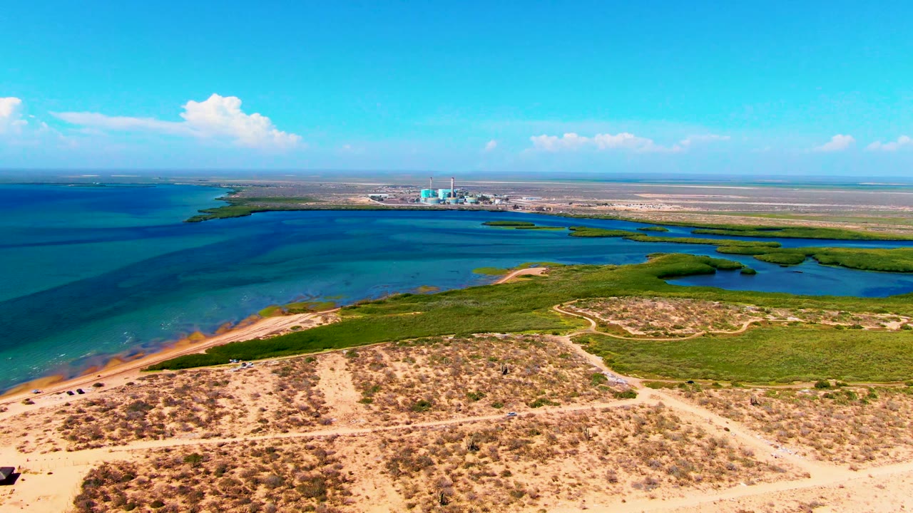 At 0:02, sandy foreground dunes and paths lead toward green wetland patches, open turquoise water, a bright cloud line, distant structures, and no visible people in San Carlos.