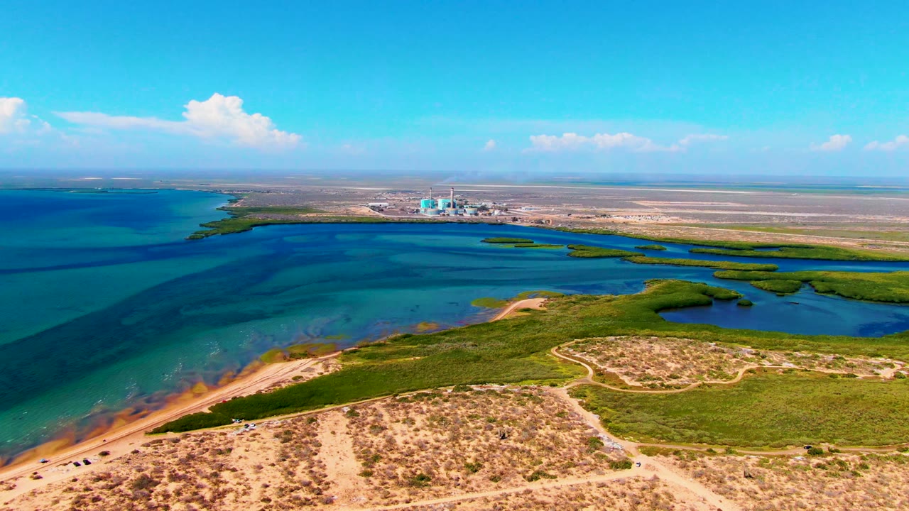 Aerial frame from San Carlos, Baja California Sur showing blue lagoon water beside sandy dunes, green wetland vegetation, a pale dirt path, distant shoreline structures, and no visible people.