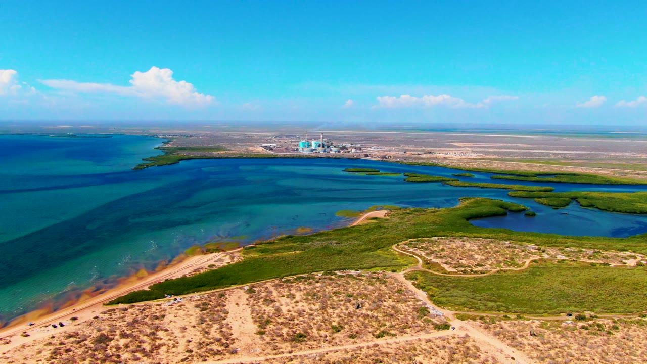 Wide aerial coastline with turquoise water, dunes, vegetation, and a small vehicle on the dirt path.