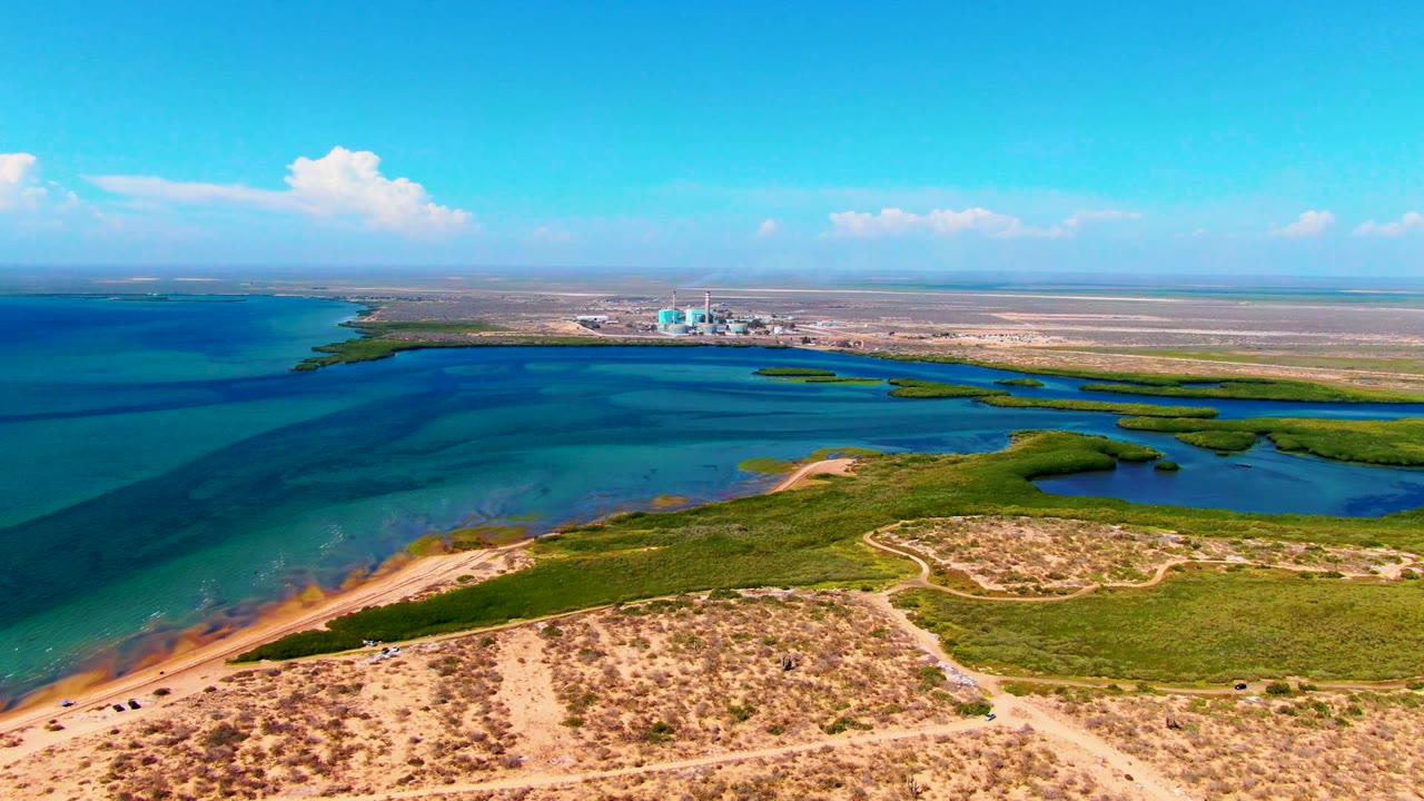 Mid-clip aerial frame from San Carlos, Baja California Sur showing the dirt path curving through sandy dunes, green vegetation edging the lagoon, distant horizon structures, and no visible people.