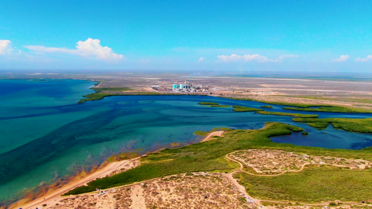 Opening frame over San Carlos, Baja California Sur shows blue-green inlet water beside tan sand, green vegetation, distant shoreline structures, bright sky, and no visible people.