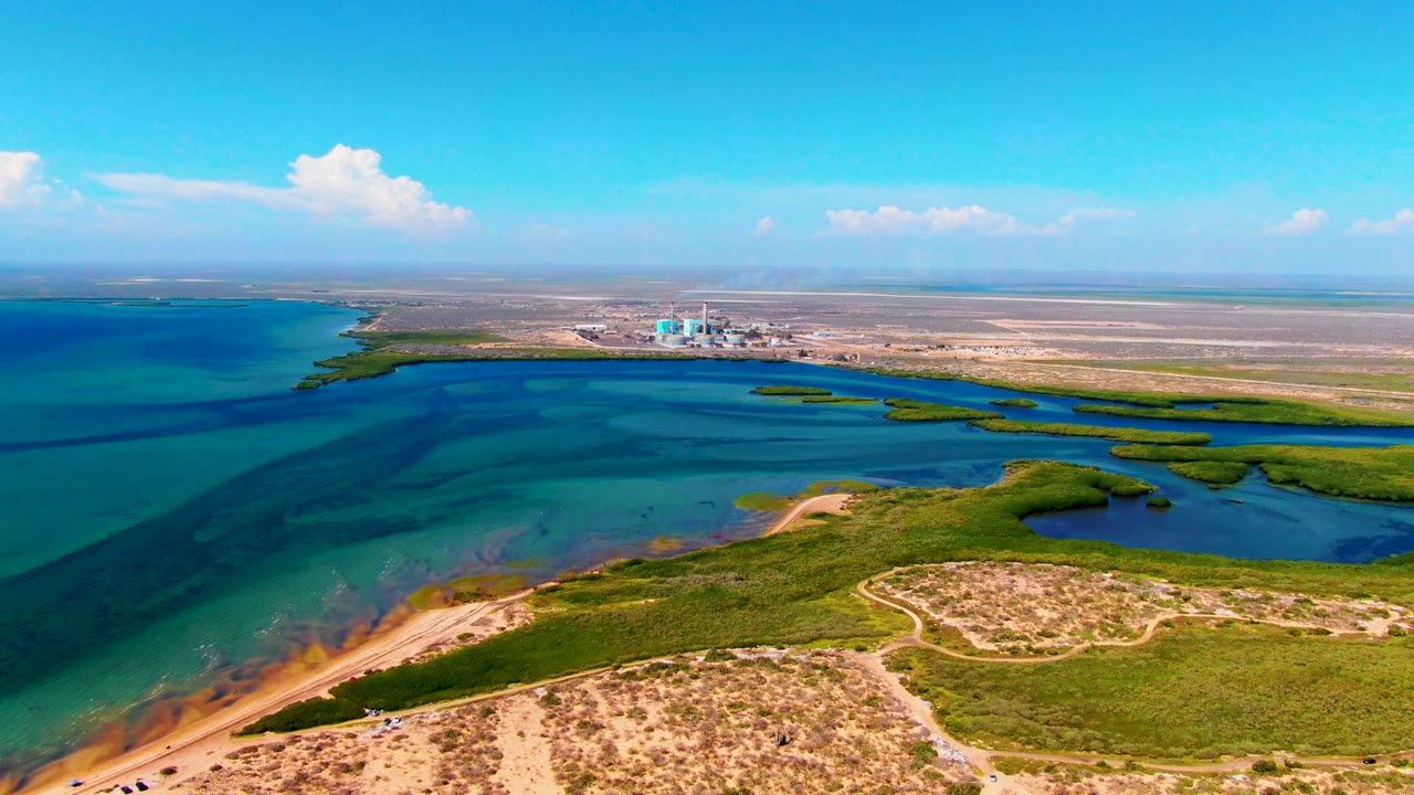Drone view across turquoise inlet water, curved sandy edge, and green coastal brush.