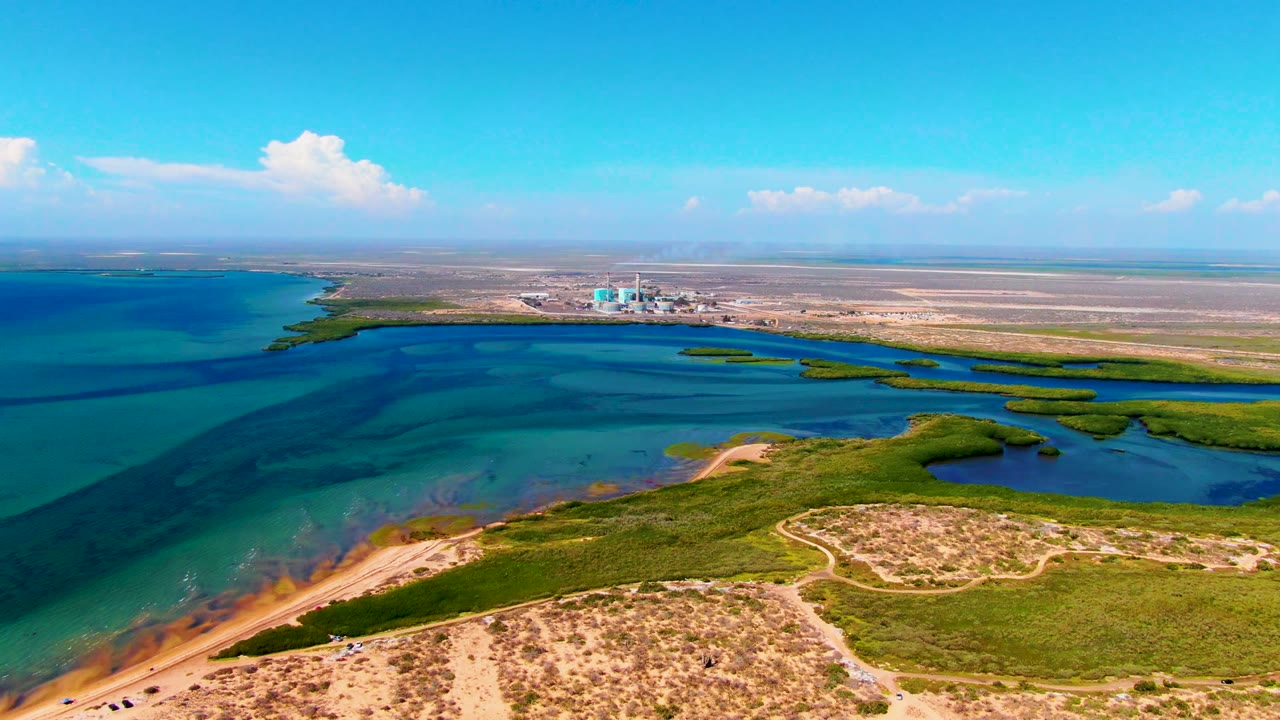 At 0:02, the drone-height frame shows shallow blue and green water spreading toward sandy flats, with distant shoreline buildings on the horizon and no visible people.