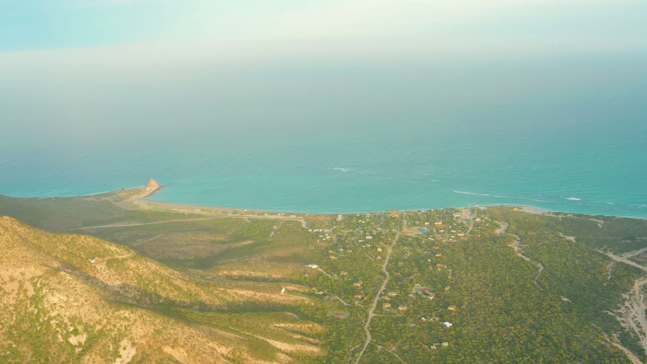 Aerial view over Cabo Pulmo, Baja California Sur, with sunlit hills in the foreground, scattered coastal buildings, winding roads, turquoise ocean, and no visible people.