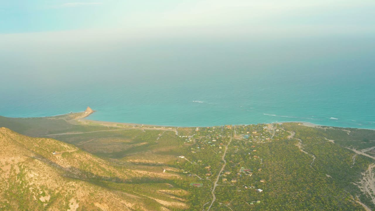 Wide aerial frame from Cabo Pulmo showing the hillside edge, a grid of vegetation and roads, scattered buildings near the shoreline, soft cloud cover, turquoise water, and no visible people.