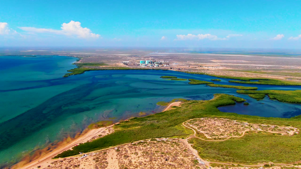 Aerial view over San Carlos, Baja California Sur, with turquoise coastal water at left, sandy dunes and green wetland patches below, distant industrial buildings on the horizon, and no visible people.