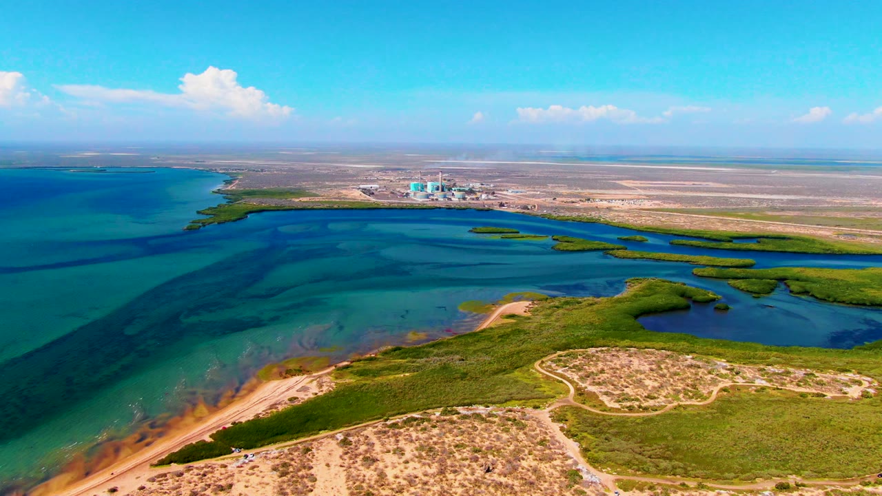 Wide aerial coastline with shallow blue water, low green islands, sandy ground, and industrial structures in the distance.
