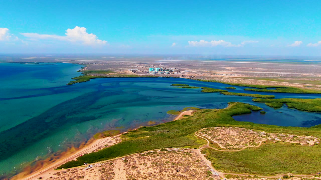Aerial view in San Carlos, Baja California Sur, with bright sandy dune areas in the foreground, blue coastal water across the middle, distant buildings near the horizon, and no visible people.