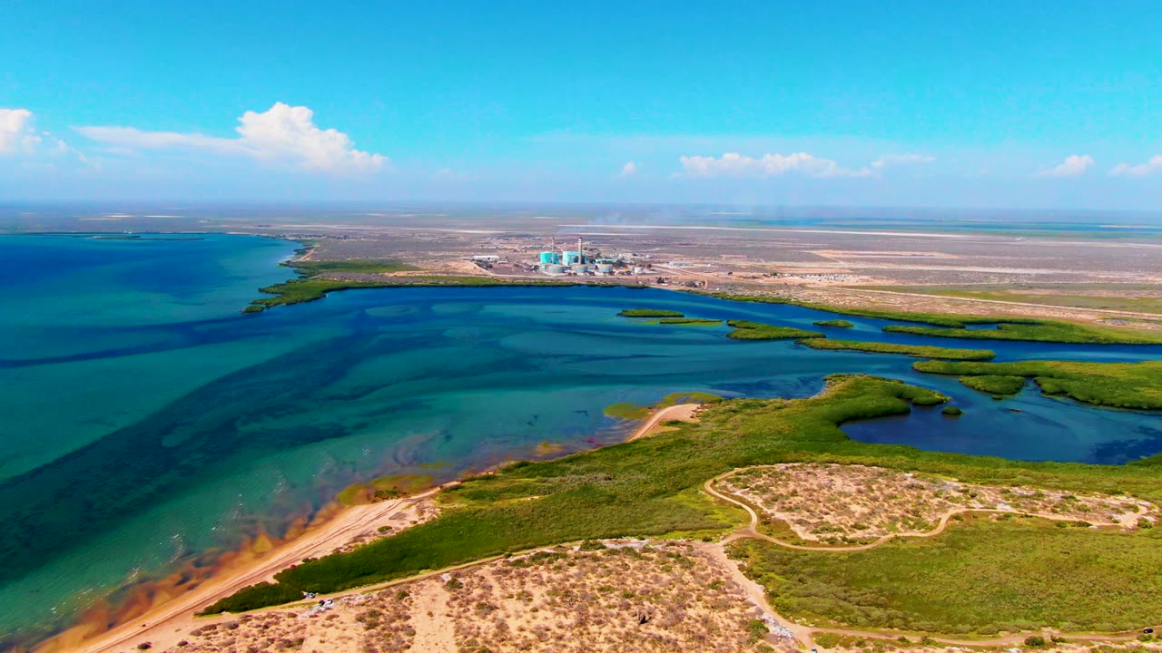 Aerial coastal scene with dunes in front, blue-green water beyond, wetland vegetation, and industry on the horizon.