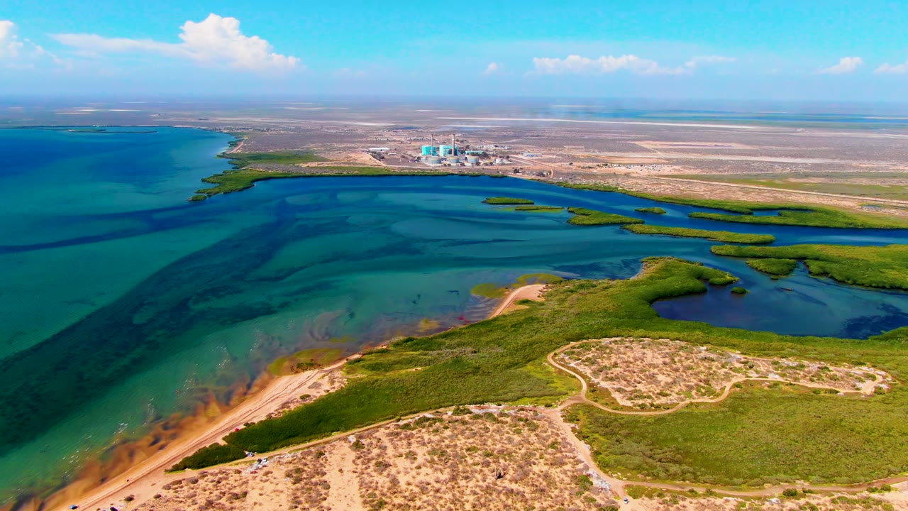 Opening aerial frame over San Carlos, Baja California Sur, with turquoise water, green wetland patches, sandy foreground tracks, distant shoreline structures, scattered clouds, and no visible people.