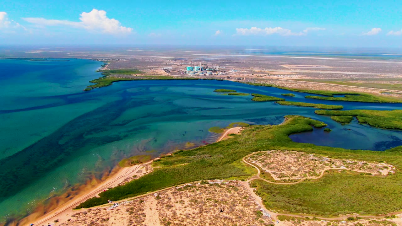 Drone view across turquoise water and marsh islands with a dark shape stretching through the bay.