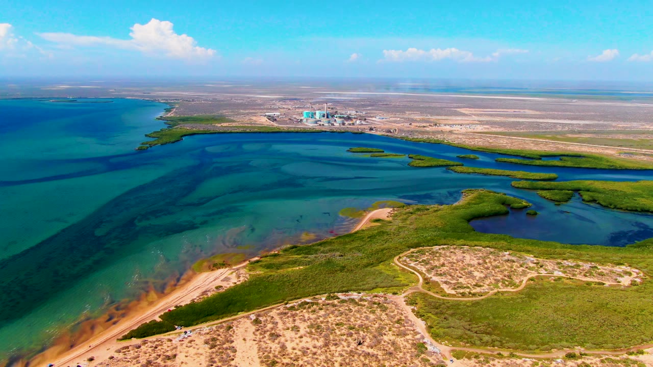 San Carlos aerial frame with the sandy foreground lower in view, green wetland channels, blue bay water crossed by a dark band, distant shore structures, clear sky, and no visible people.