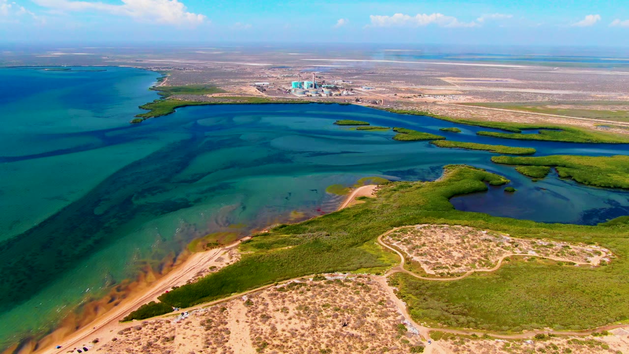 Aerial view of San Carlos, Baja California Sur coastline with turquoise water, a sandy foreground beach, green shoreline vegetation, distant open land, and a small person visible near the lower beach area.