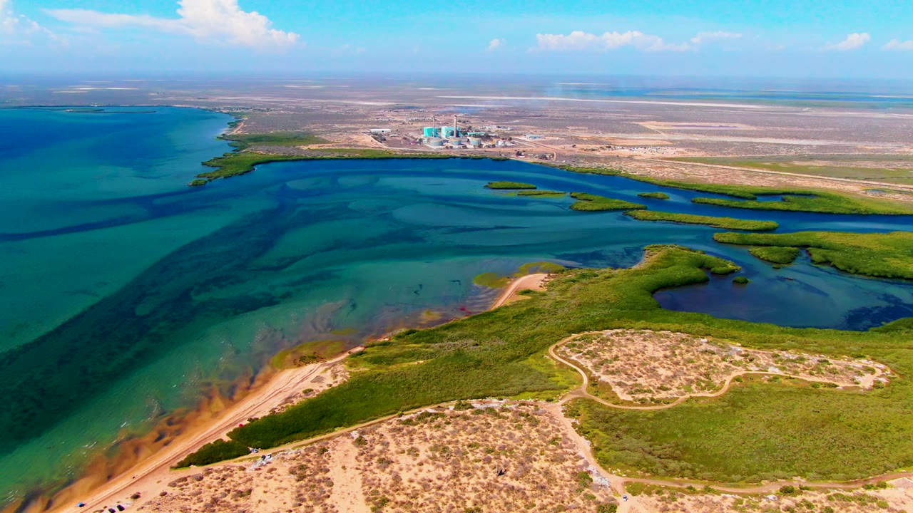 San Carlos, Baja California Sur aerial frame with bright blue water across the left and center, green vegetation along the shore, sandy paths in the foreground, distant structures, and a small beach figure still visible.