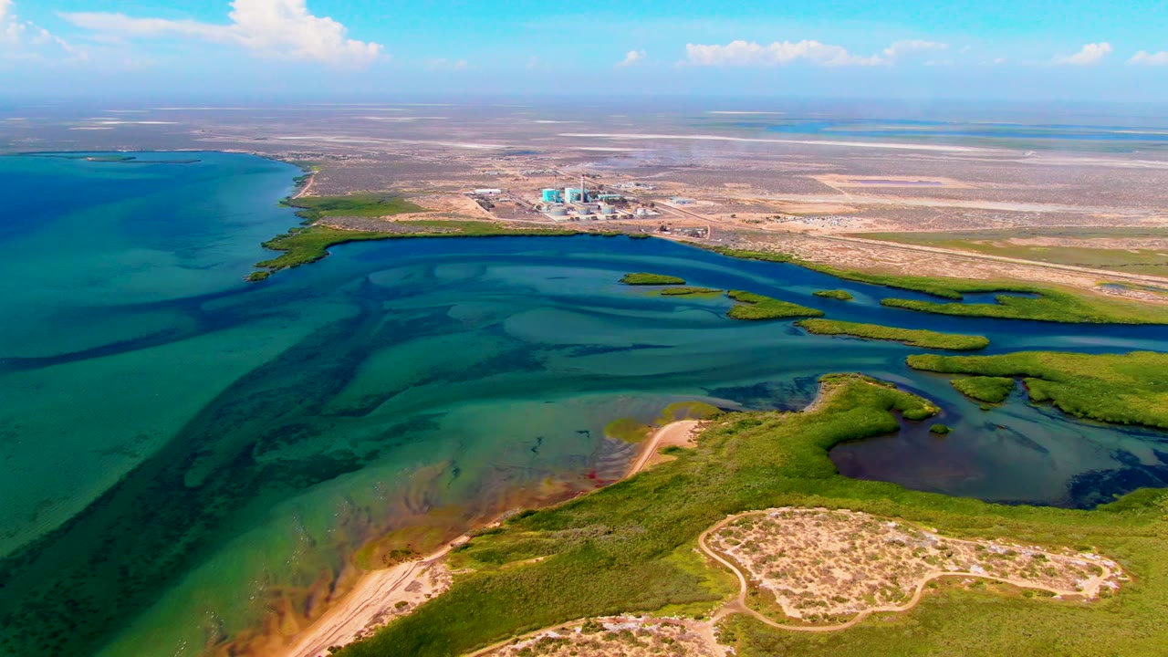 Aerial frame over San Carlos, Baja California Sur, showing turquoise water channels, green marsh patches, sandy foreground land, a distant blue-roofed structure, and no visible people.