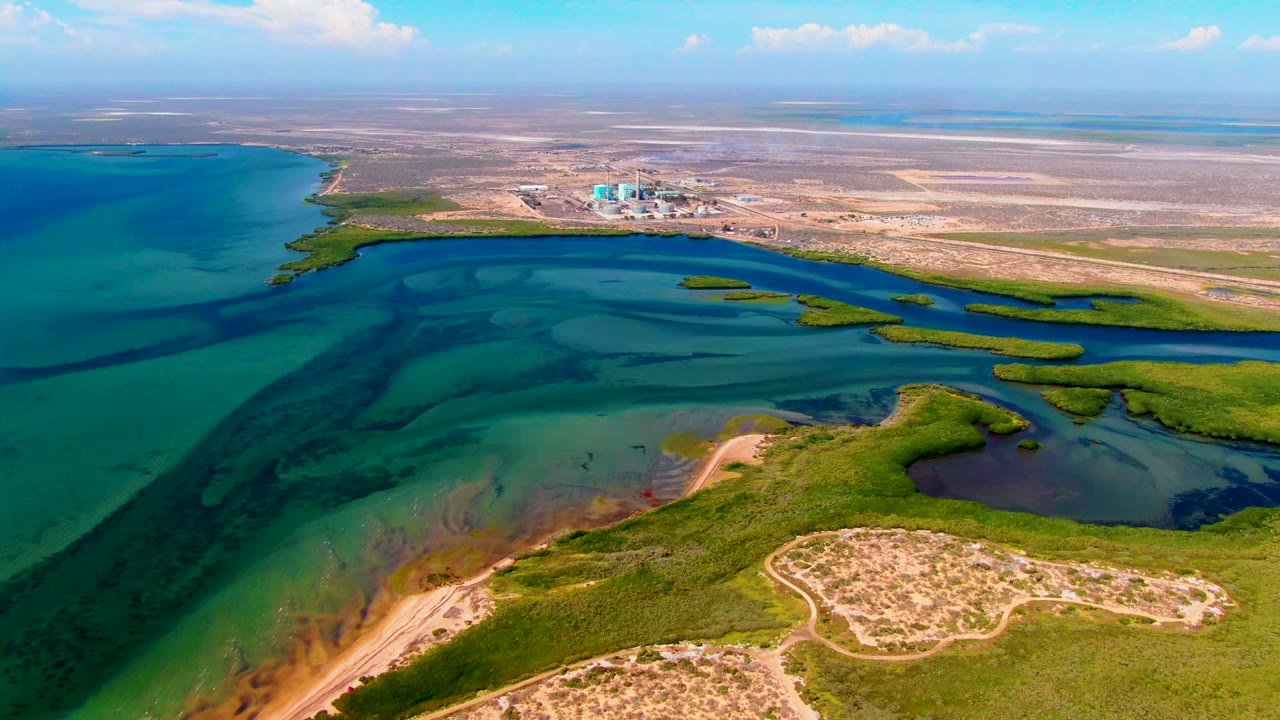 At 0:02, the drone-height view shows San Carlos wetlands with a green shoreline band, dark blue water channels, turquoise shallows, pale dry terrain beyond, and no visible people.