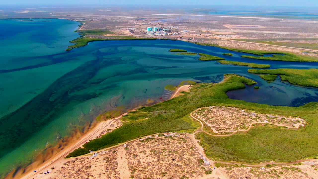 Opening aerial frame over San Carlos, Baja California Sur, showing turquoise lagoon water, sandy dune foreground, green marsh patches, distant shore structures, and no visible people.