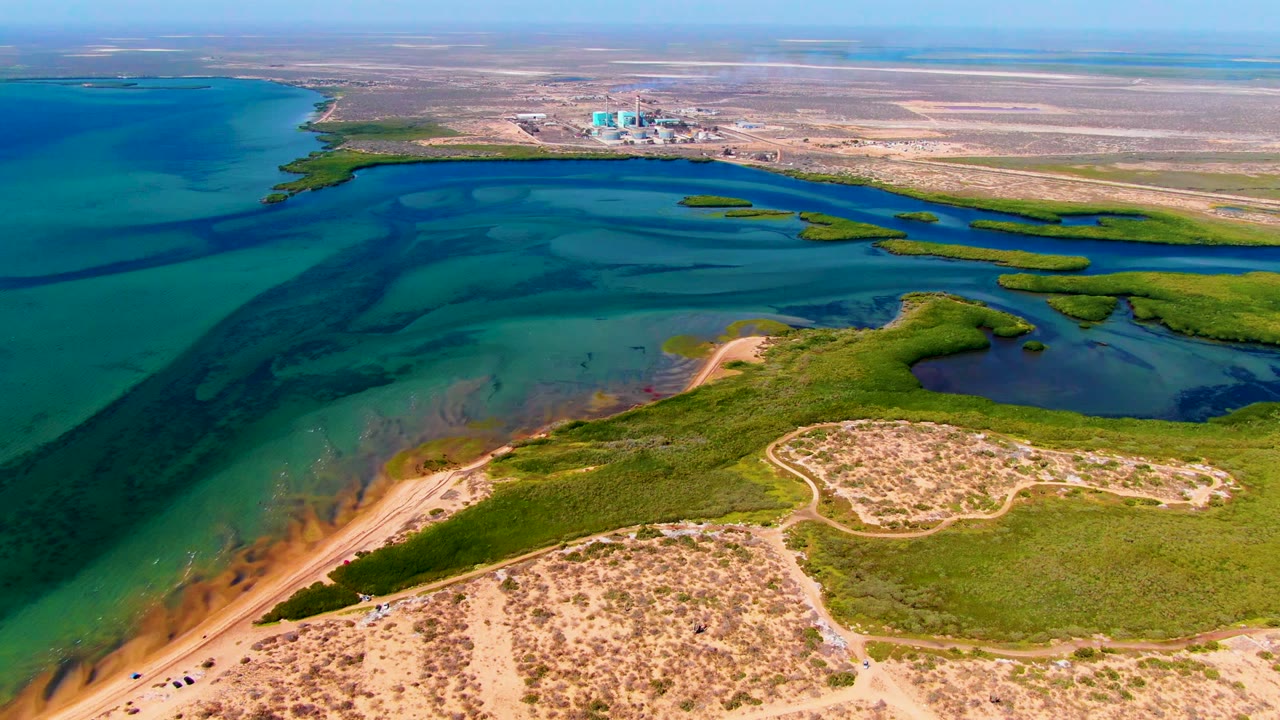 Aerial frame of lagoon water, marshland, sandy dunes, distant buildings, and flat dry land.