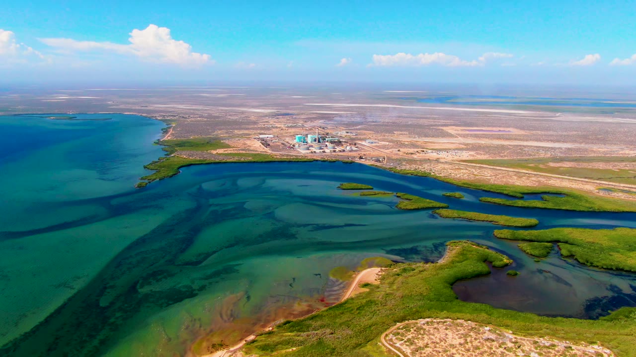Opening aerial frame over San Carlos, Baja California Sur, showing turquoise wetland water, green marsh edges, a pale sandbar, dry inland flats, distant structures, scattered clouds, and no visible people.