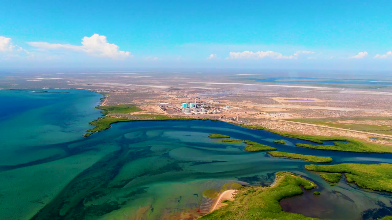 San Carlos aerial frame showing broad blue-green water on the left, narrow marsh channels across the center, dry terrain beyond, scattered clouds, and no visible people.