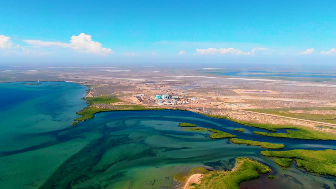 Aerial coastal wetland scene with turquoise water, green marsh islands, and a dry inland background.