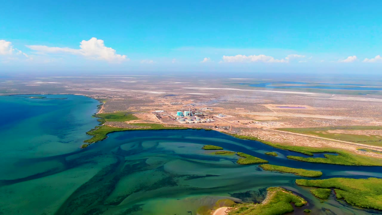 Opening aerial frame over San Carlos, Baja California Sur, with turquoise shallows in the foreground, sandy shoreline and vegetation beyond, distant structures inland, and no visible people.