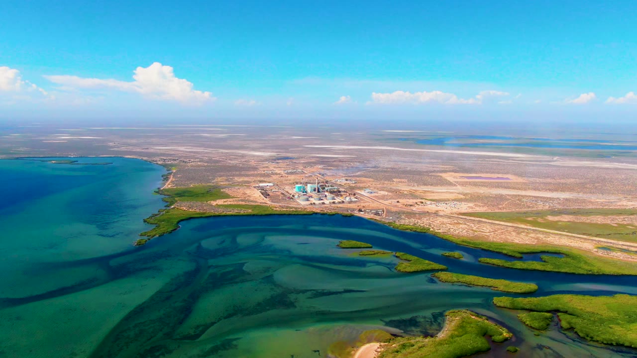 Aerial frame at 0:01 over San Carlos shows the inlet curving across blue and green water, low vegetation on the shore, pale sandy ground inland, and no visible people.
