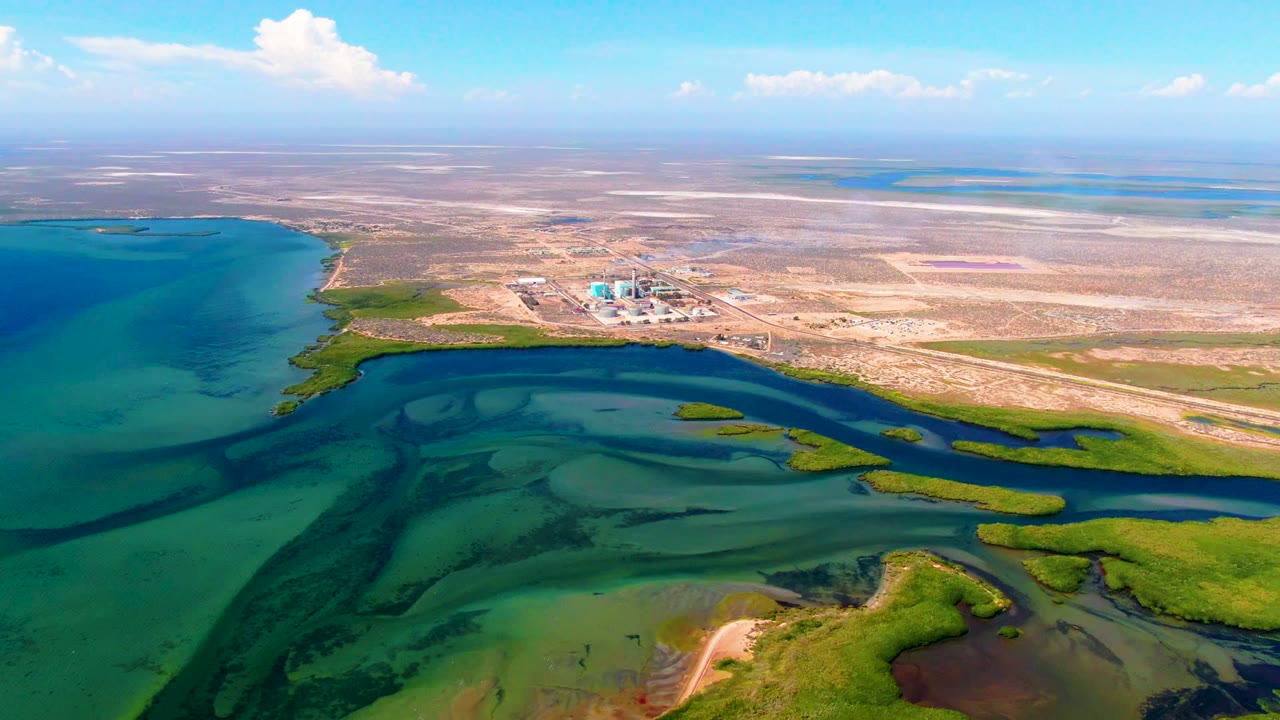 High aerial view over San Carlos, Baja California Sur, showing turquoise wetland water channels, green marsh patches, dry flats, a small distant structure, blue sky, and no visible people.