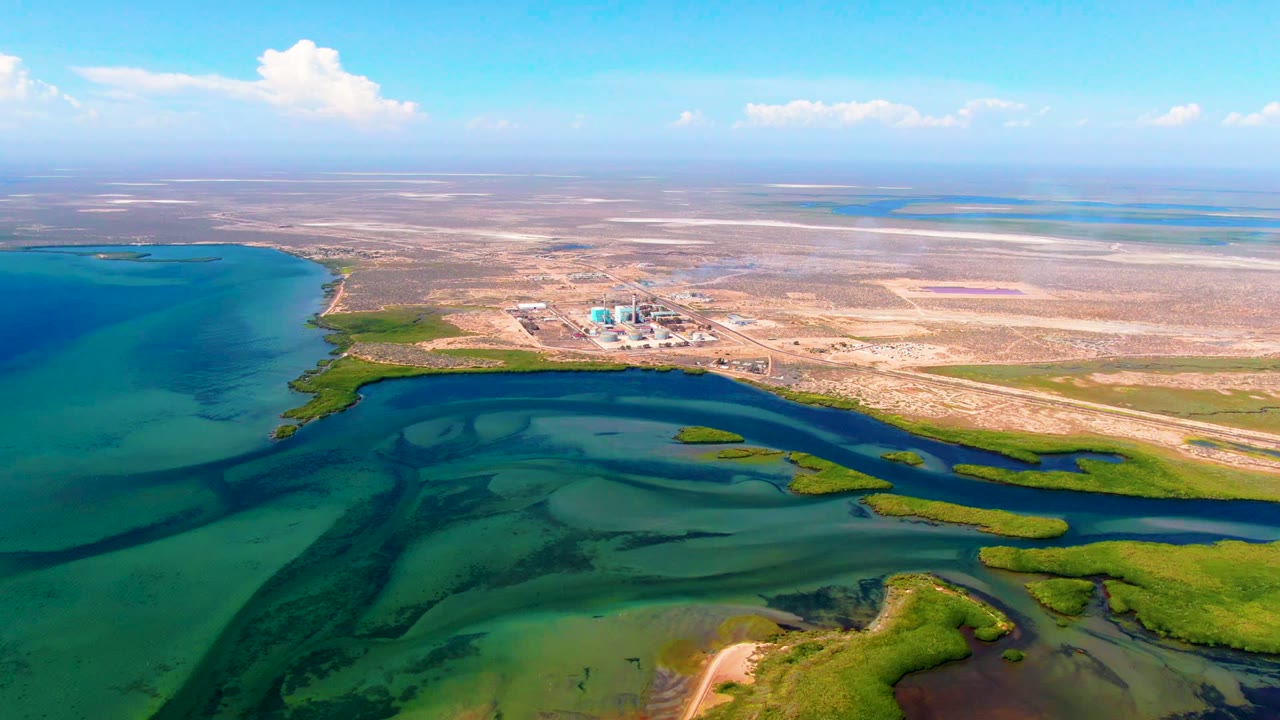 High oblique aerial over San Carlos, Baja California Sur, showing dark winding channels through shallow turquoise water, green marsh vegetation, small distant buildings, dry flats, and no visible people.