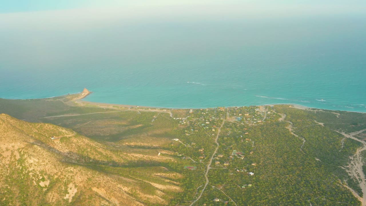 Aerial view over Cabo Pulmo with brown-green hillside in the foreground, scattered buildings and dirt roads below, blue ocean beyond, and no visible people.