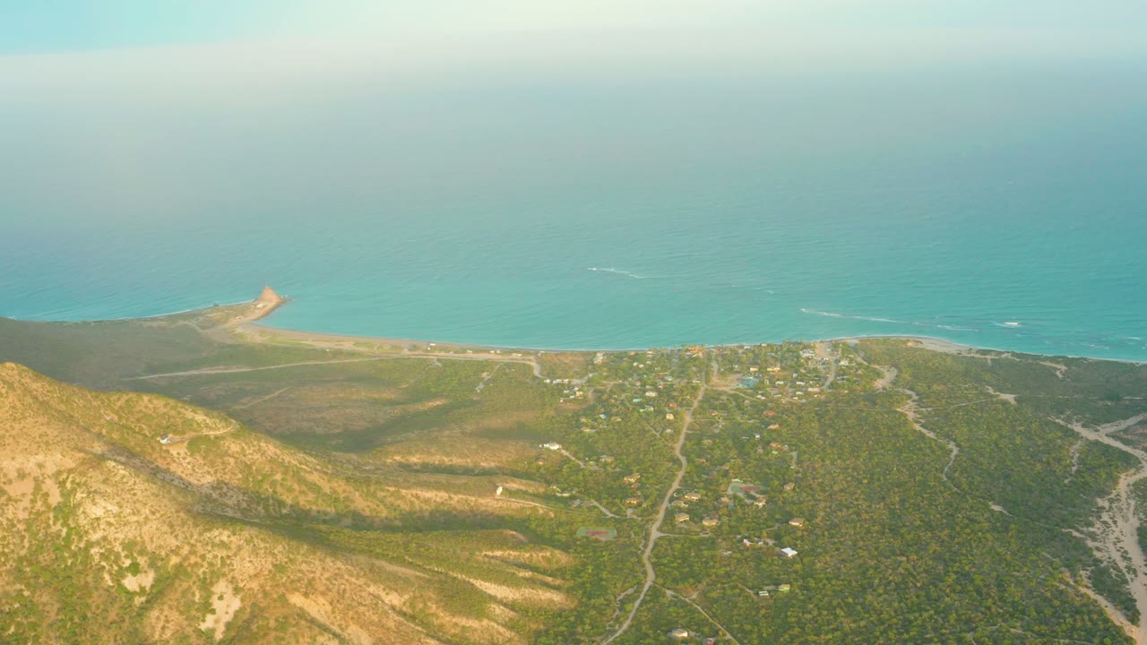 Aerial view of Cabo Pulmo with the hillside shadow pattern shifting slightly across green-brown terrain, scattered buildings, shoreline, ocean, and no visible people.