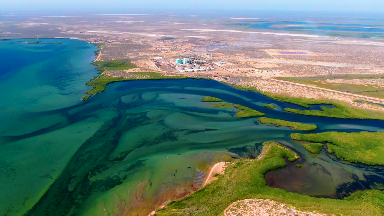 Wide aerial frame over San Carlos, Baja California Sur showing turquoise coastal water, green marsh vegetation, a pale inland industrial site, distant lagoons, and no visible people.