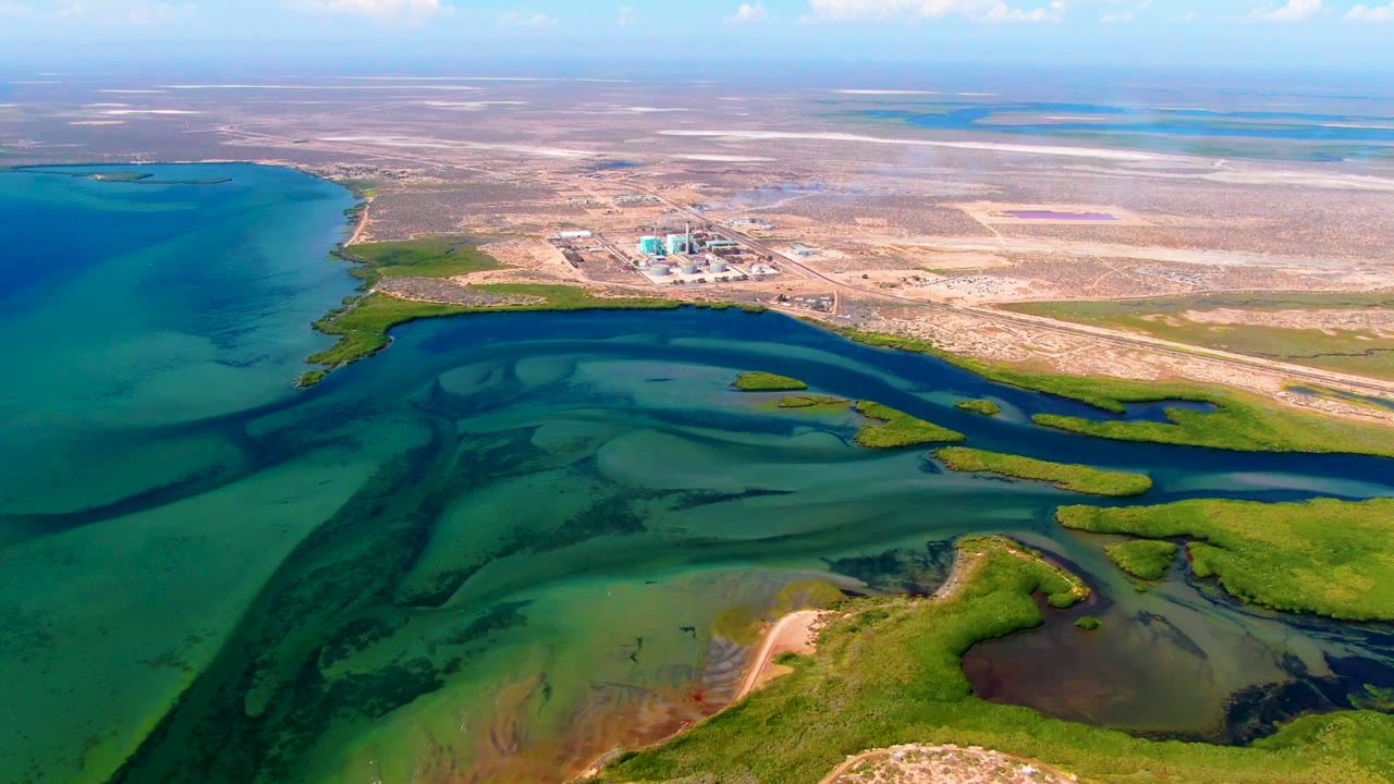 Aerial coastline with marsh channels, shallow water, dry land, and low industrial buildings.