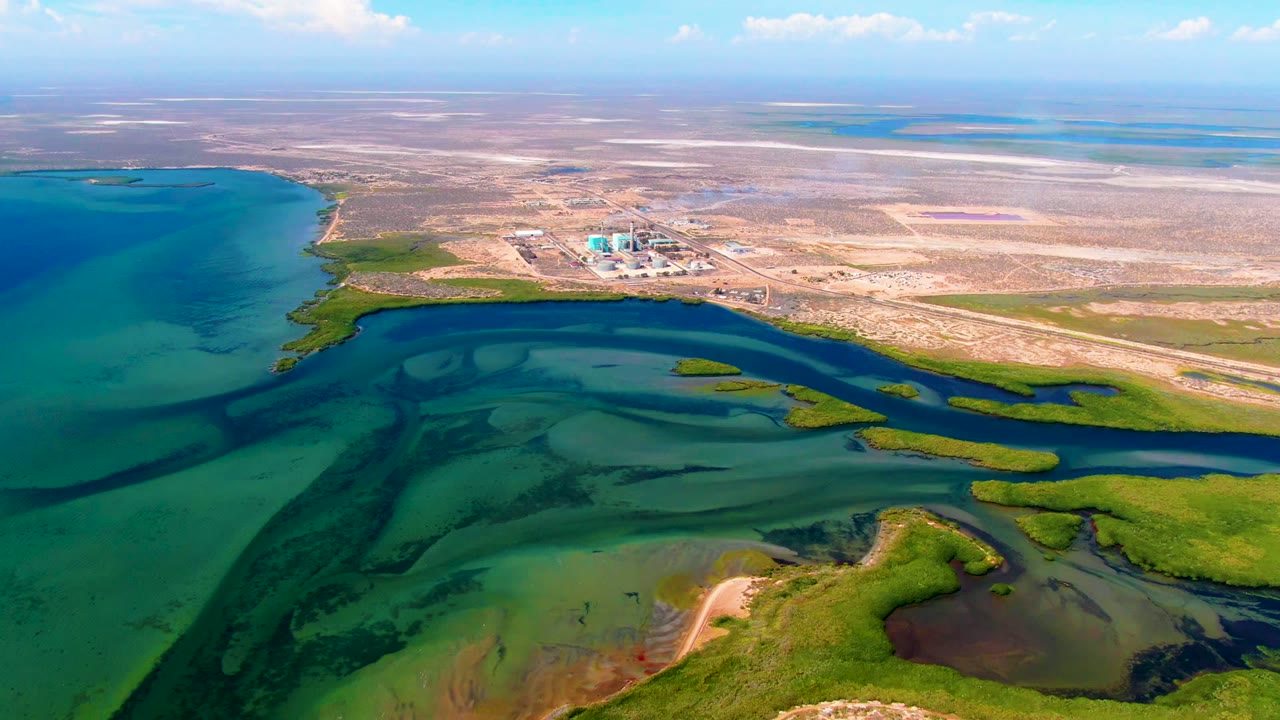 San Carlos, Baja California Sur aerial frame showing a sweeping wetland edge, dark blue channel water, green marsh patches, pale arid ground, industrial buildings, and no visible people.