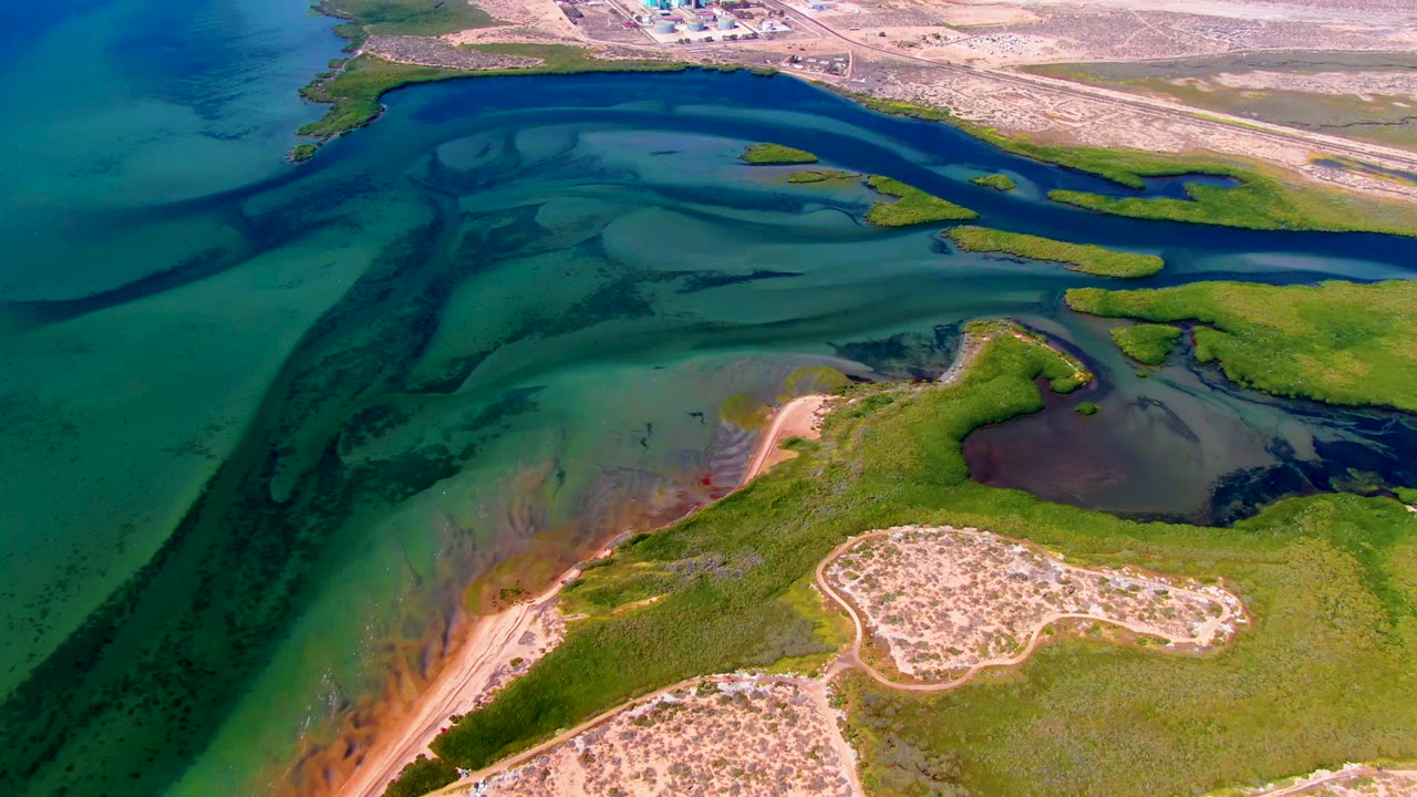 Wide aerial view of shallow coastal channels, marsh islands, sandy ground, and distant buildings.