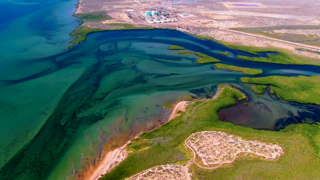 Aerial San Carlos frame with broad turquoise water at left, dark curving tidal channels, green marshland, pale sand, and a cluster of distant structures, with no visible people.